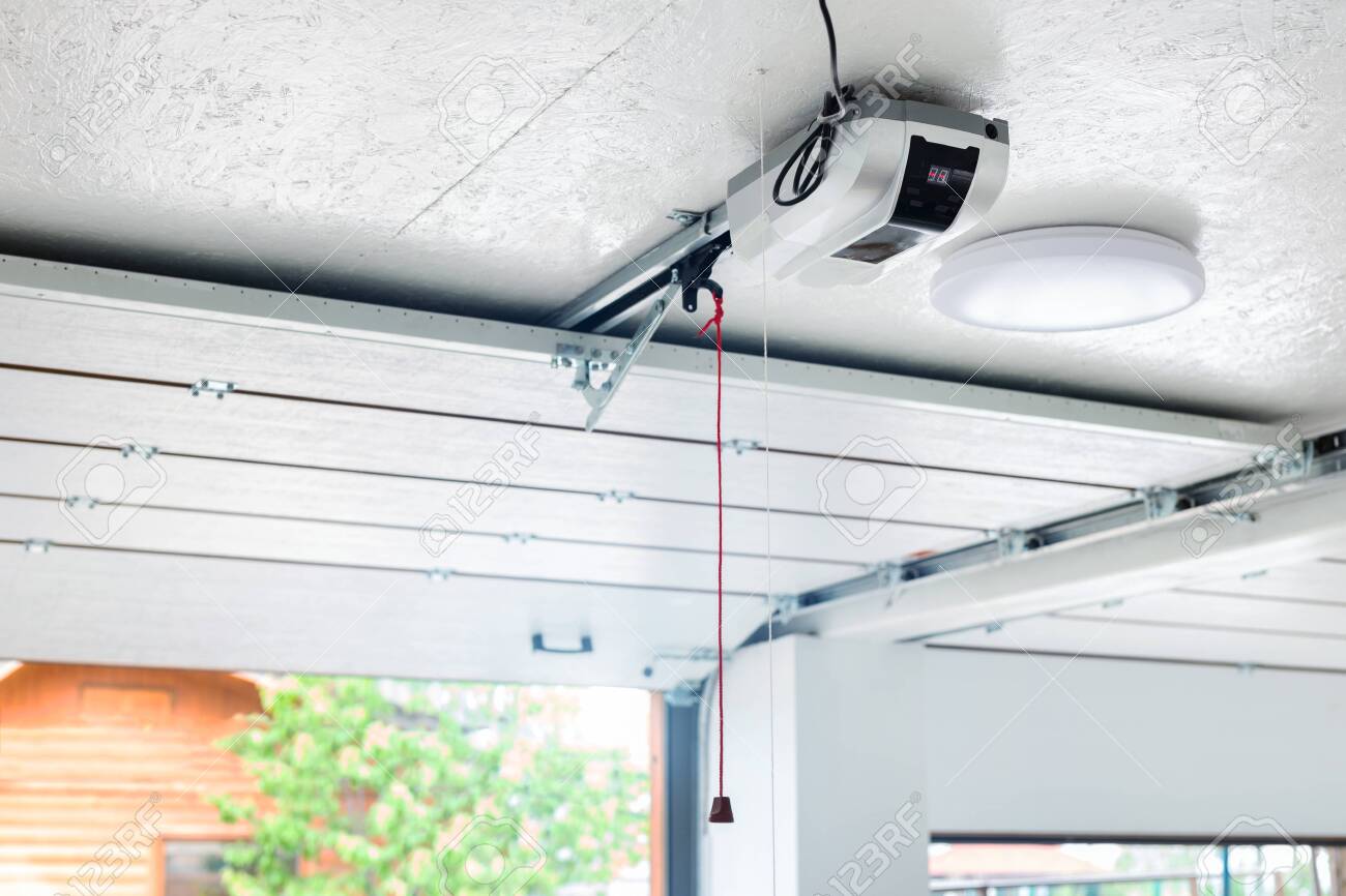 Opening door and automatic garage door opener electric engine gear mounted on ceiling with emergency cord. Double place empty garage interior with rolling entrance gate.